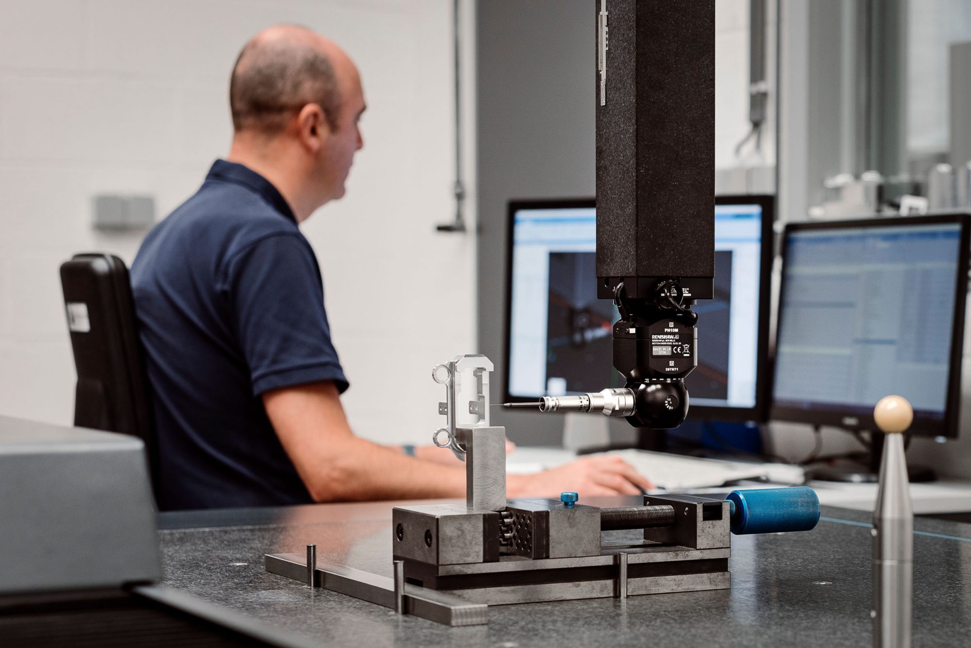 Machine à mesurer tridimensionnelle avec un marbre et un étau dans lequel est serré un composant en aluminium. Le capteur de mesure se situe à droite du composant. Le responsable AQ travaillant sur son ordinateur est visible à l‘arrière-plan.