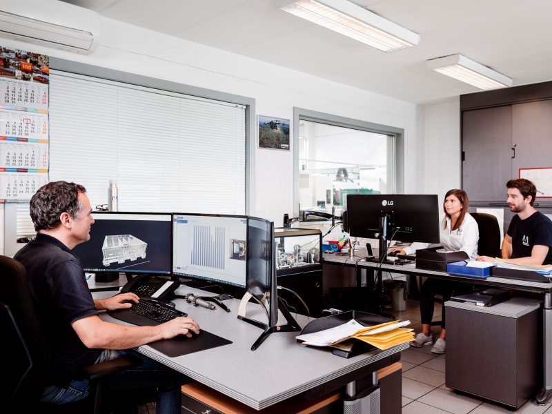 The COO sits in front of three computer screens. Opposite him at another desk, the purchasing manager and another Mockel employee sit in front of a screen.