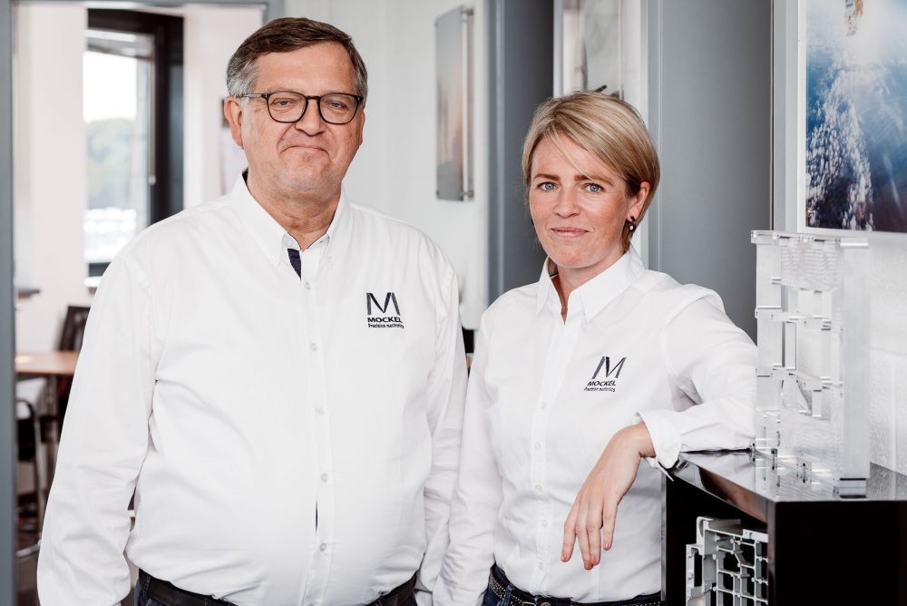 mockel-owner-and-ceo-with-aluminium-casing-looking-into-camera-1 In die Kamera blicken Firmeninhaber Robert Mockel und Geschäftsführerin Annabelle Mockel, ihr Arm liegt auf einem Regal mit Aluminiumgehäusen
