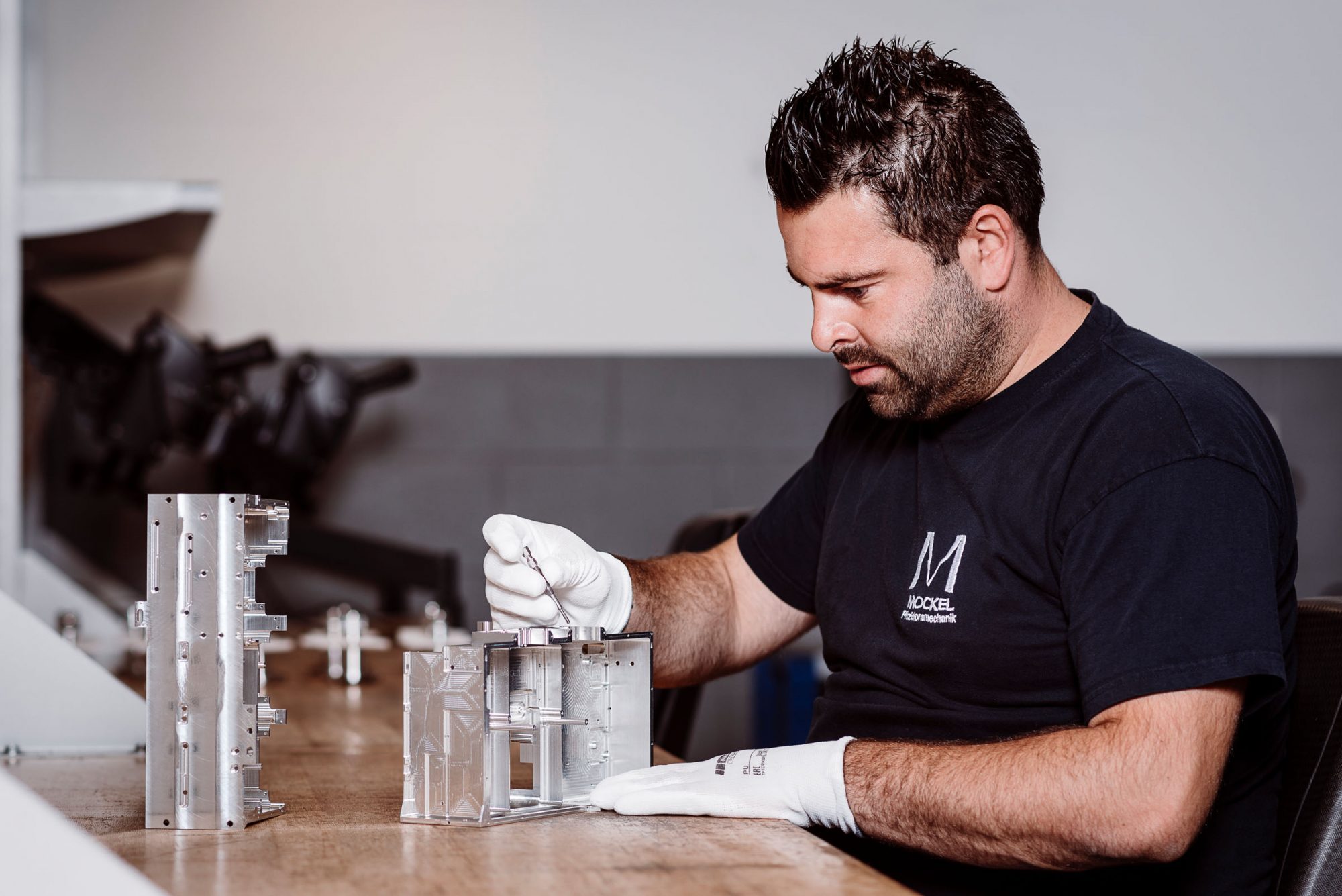 An assembly worker secures an aluminium housing with one hand while holding a tool with the other. He is wearing white gloves.